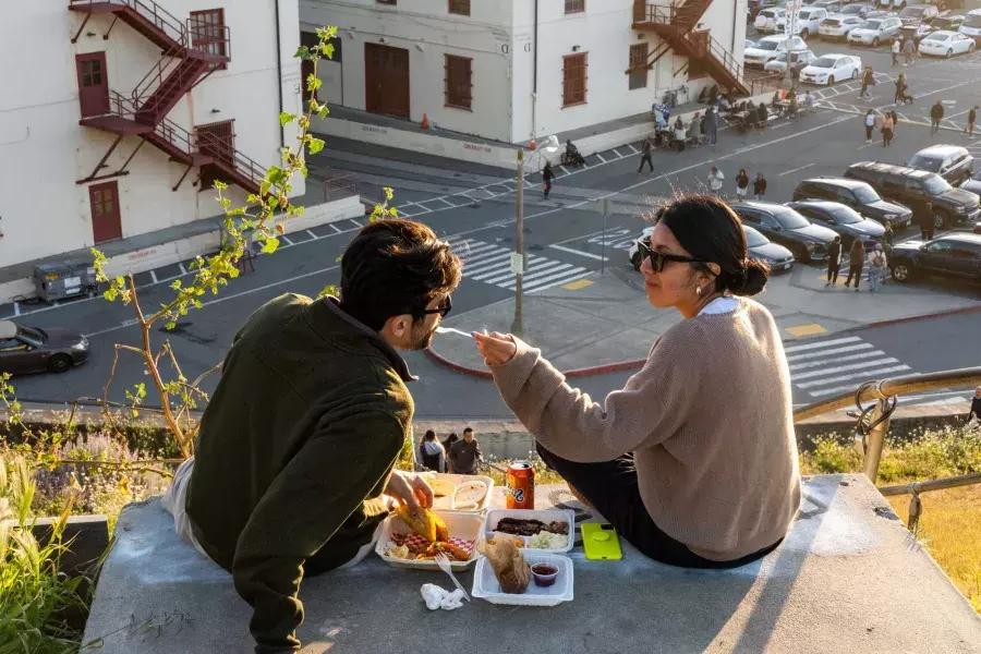 샌프란시스코의 포트메이슨 센터에서 한 커플이 야외 식사를 하고 있습니다. 여자는 동반자에게 음식 맛을 먹여줍니다.