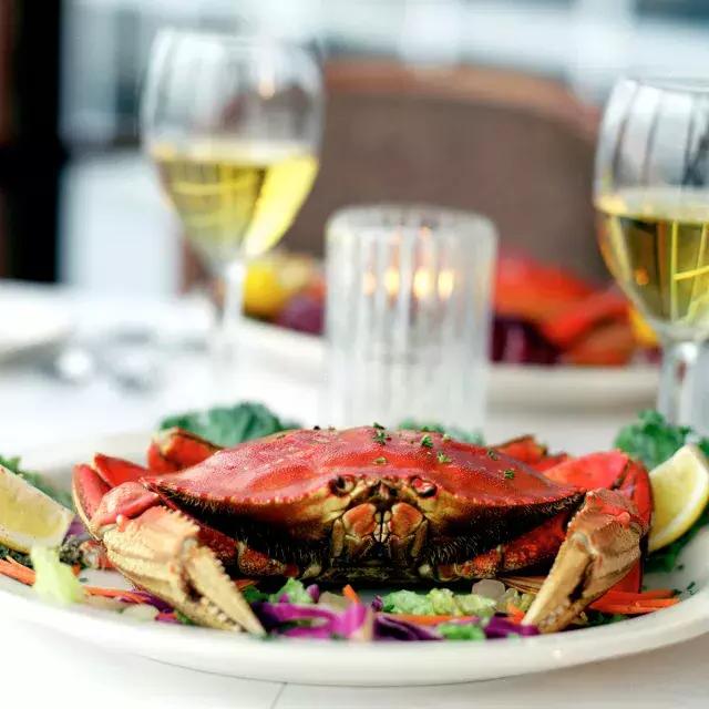 A Dungeness crab sits on a plate in a 餐厅 with two glasses of white wine in the background.
