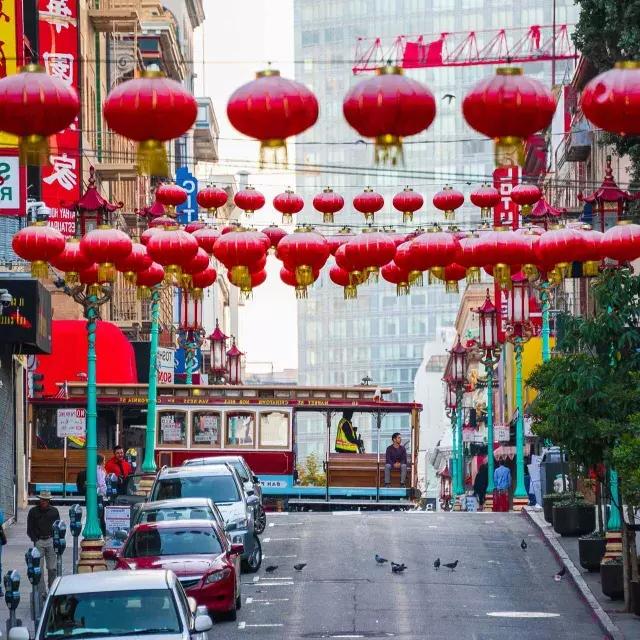 Une rue vallonnée du quartier chinois de 贝博体彩app est représentée avec des lanternes rouges suspendues et un tramway qui passe.